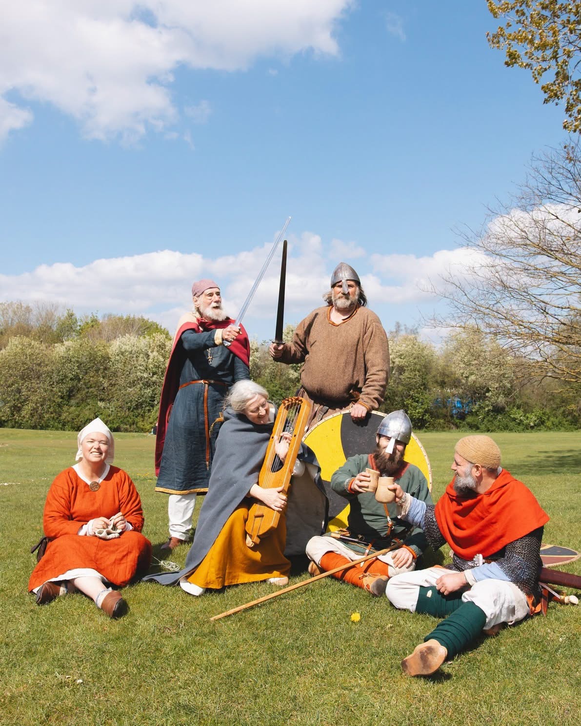 A group of Viking performers sit on a lawn with trees in the background 
